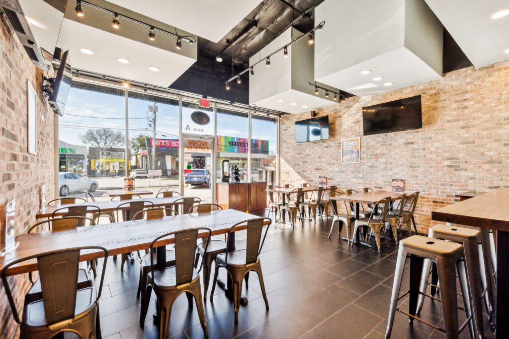 Interior of a restaurant with brick walls, metal chairs, and wooden tables, featuring floor-to-ceiling windows looking out onto a street.