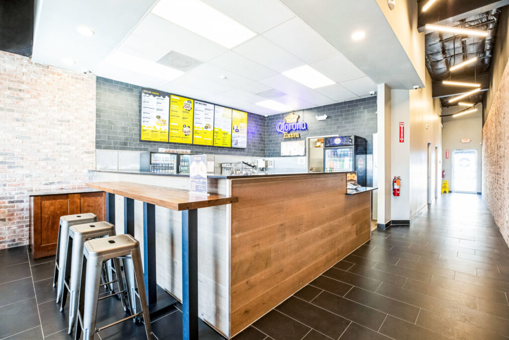 A wooden service counter in a cafe with a menu board on the wall, a Corona Extra sign, and metal bar stools in the foreground.