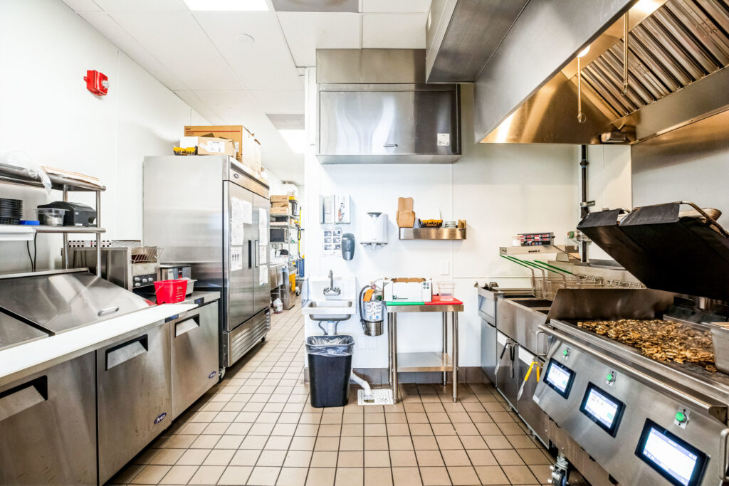 A commercial kitchen with stainless steel appliances, including a reach-in refrigerator, a flat-top grill with food, a small sink, and tiled floors.