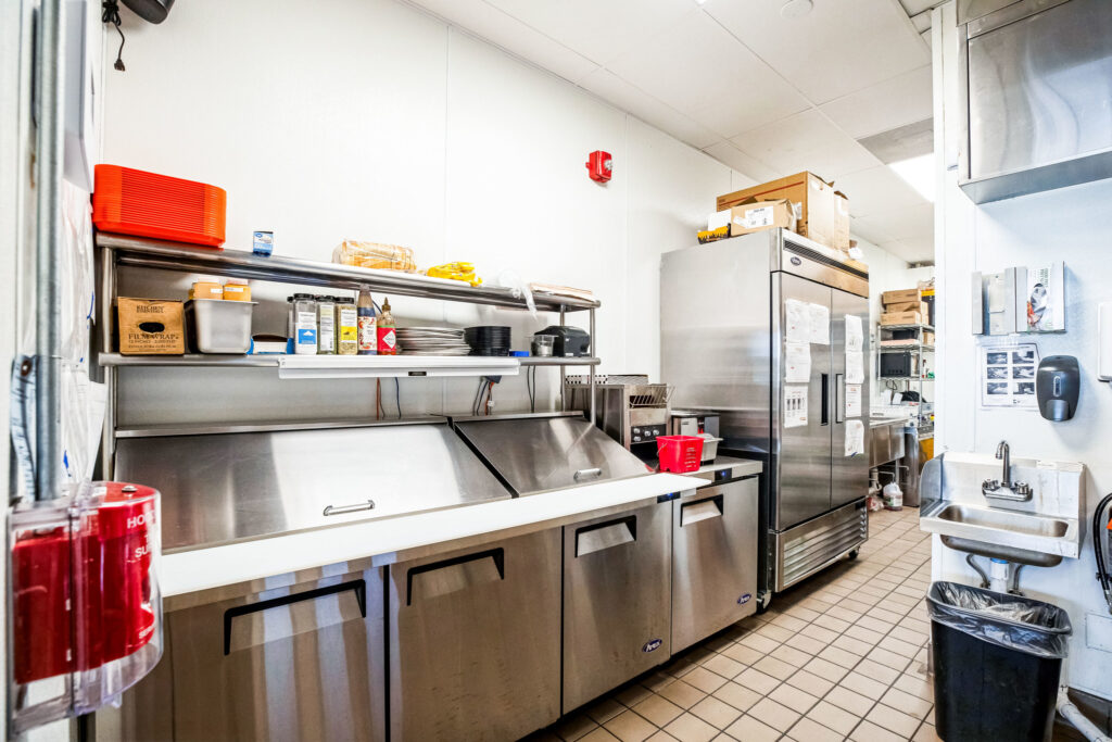 Commercial kitchen featuring stainless steel prep tables, a large reach-in refrigerator, a wall-mounted sink, and shelves stocked with kitchen supplies and food containers.