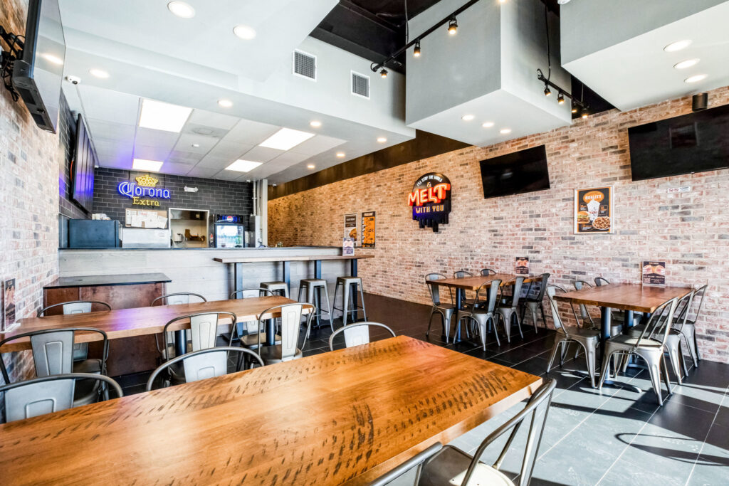 Interior of a restaurant with brick walls, wooden tables, metal chairs, a bar counter, and neon signs.
