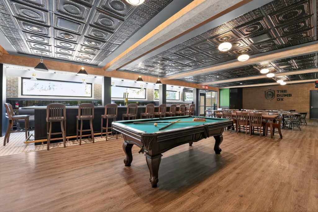A billiard table in the center of a restaurant with a bar featuring high stools and a dining area with wooden tables and chairs.