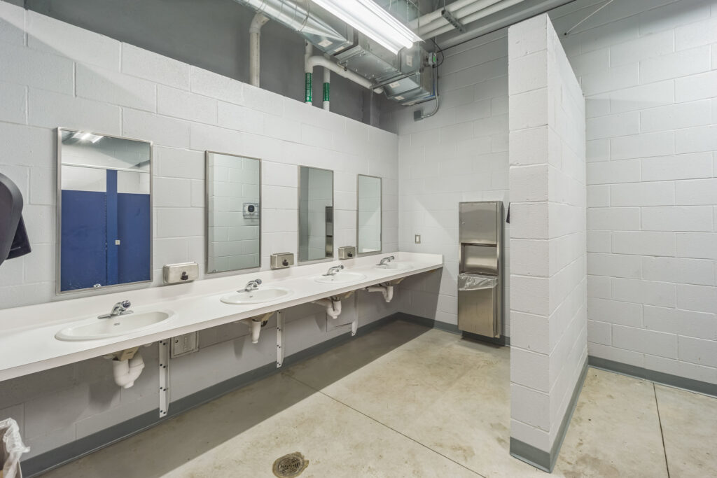 A row of three sinks with mirrors against a white block wall, next to a wall-mounted trash receptacle.