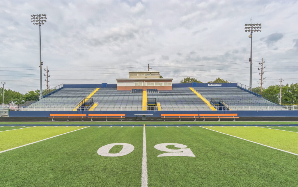 A view of an empty football stadium from the 50-yard line, showing the field and tiered bleacher seating.