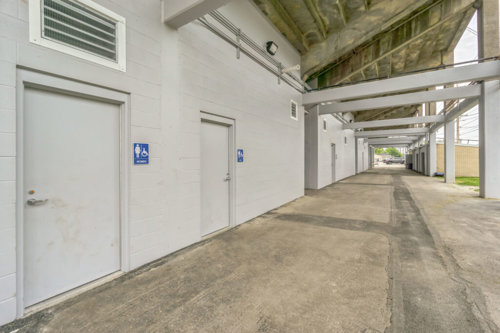 Outdoor hallway under a concrete stadium structure with doors labeled for women’s and men’s restrooms.