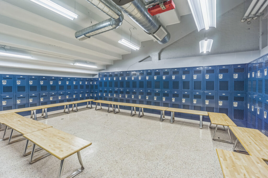A locker room featuring rows of blue metal lockers lining the walls with wooden benches positioned in front of them.