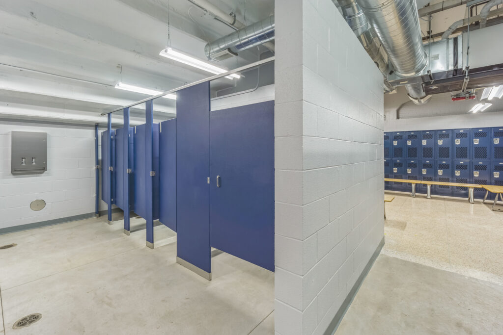 A row of blue bathroom stalls next to a block wall, with a room of blue metal lockers visible in the background.