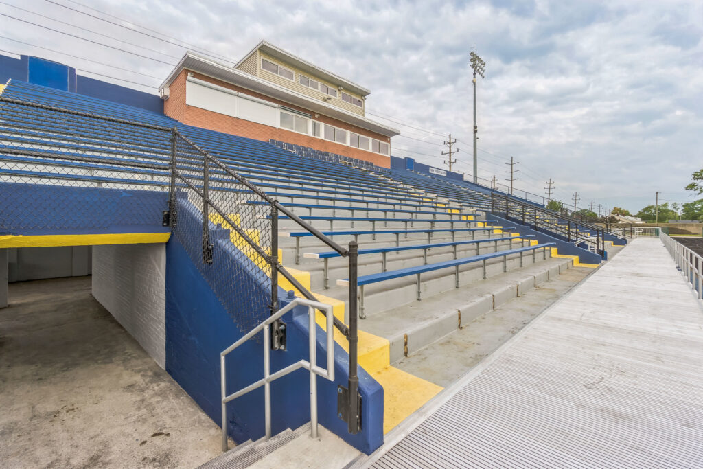 Empty outdoor stadium bleachers with blue and yellow seating, an access tunnel, and a press box.