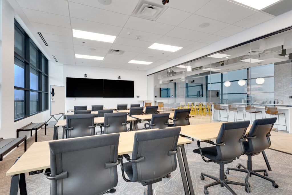 Modern office training room with rows of black chairs facing a large screen and a glass-walled kitchen area in the back.