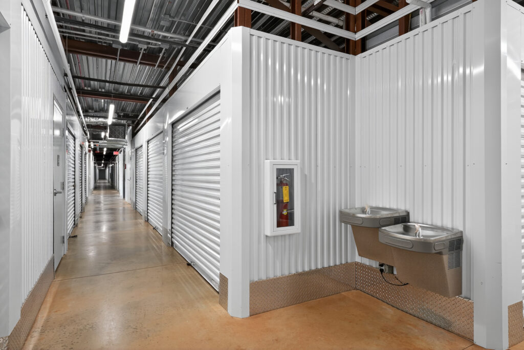 A storage facility hallway lined with white roll-up doors, featuring a wall-mounted fire extinguisher cabinet and two drinking fountains.