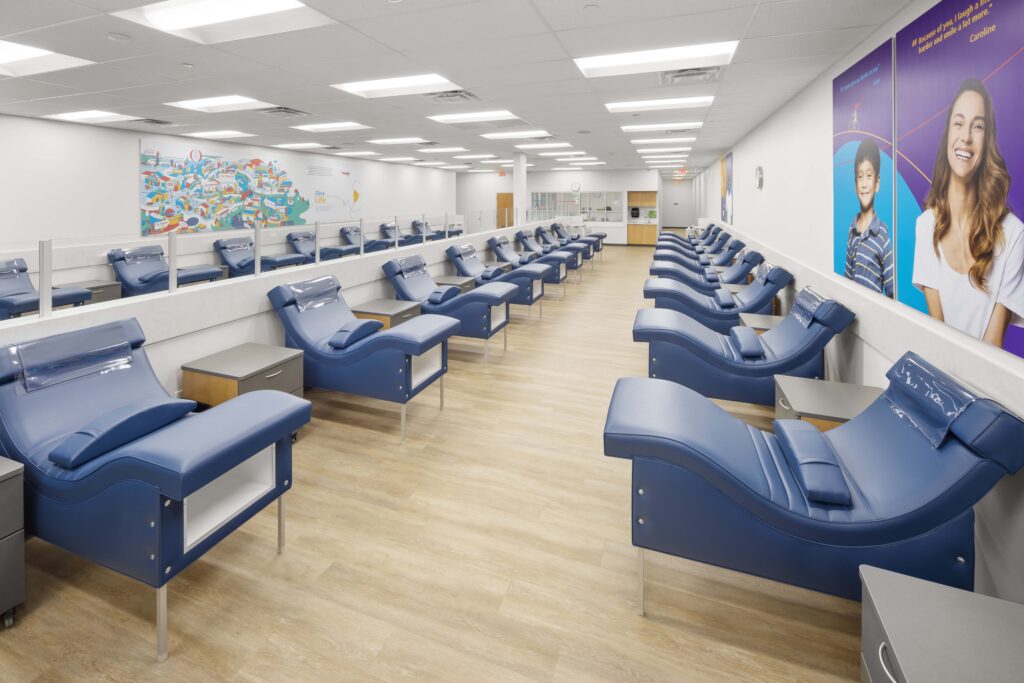 A blood donation center with rows of blue reclining chairs facing each other across a central aisle.