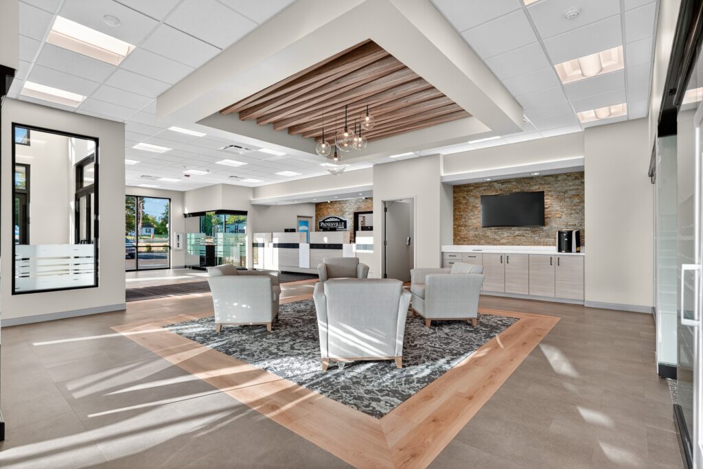 A hotel lobby with three gray armchairs arranged around a patterned rug, a reception desk in the back, and a television mounted on a stone wall.