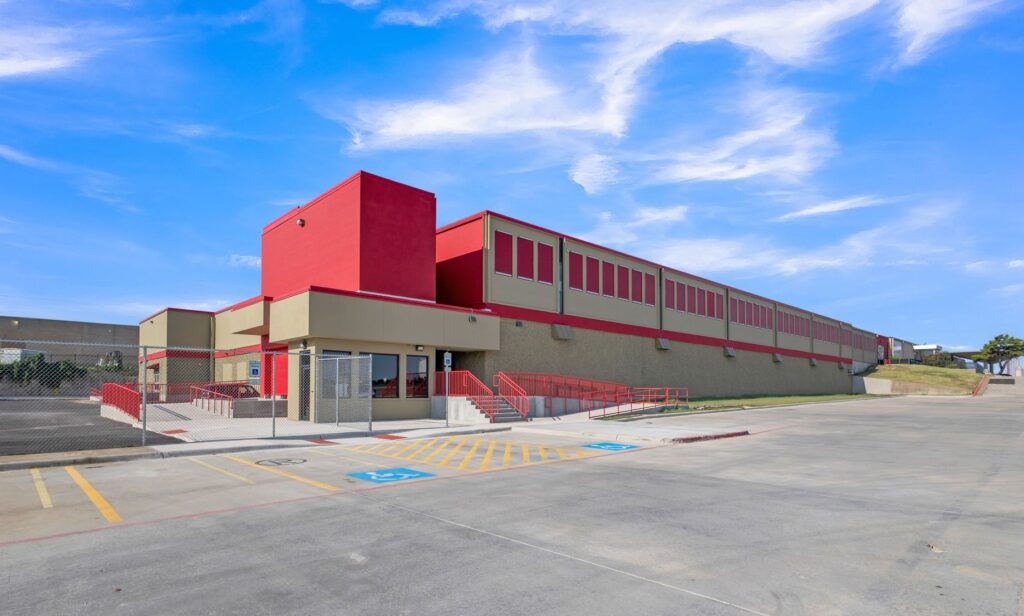 Modern school building with red and tan exterior, featuring a ramp and stairs leading to the main entrance and a marked accessible parking space.
