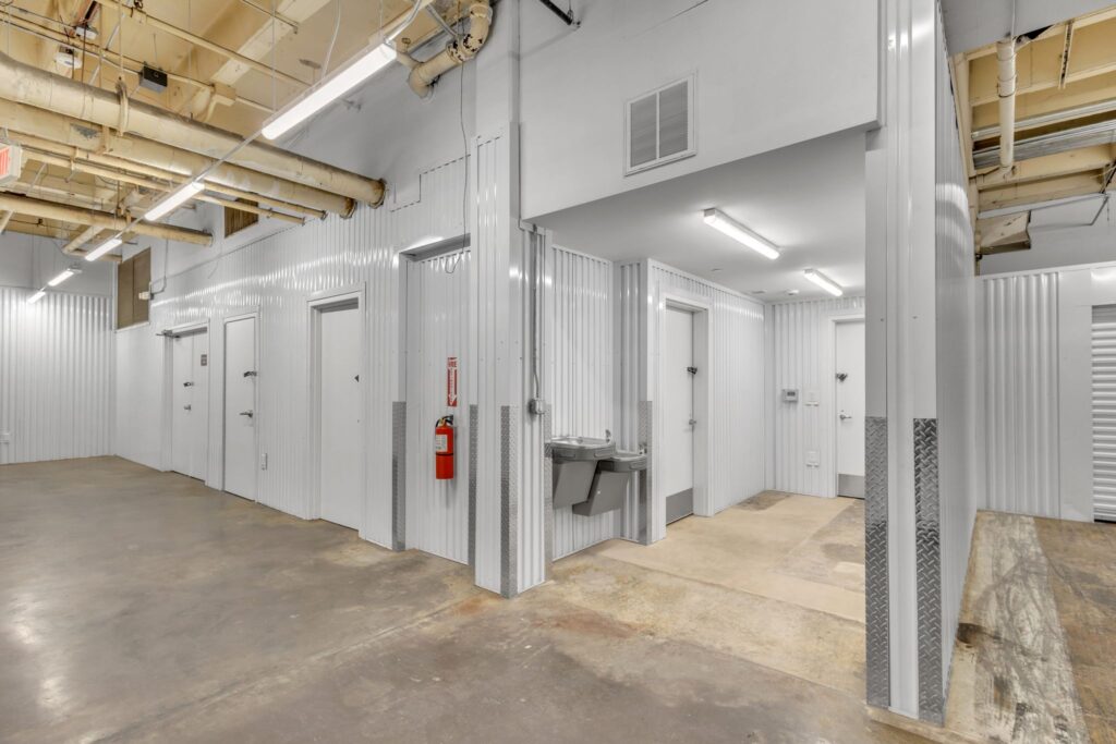 Storage unit hallway with white metal walls, multiple unit doors, a fire extinguisher, and a wall-mounted drinking fountain.
