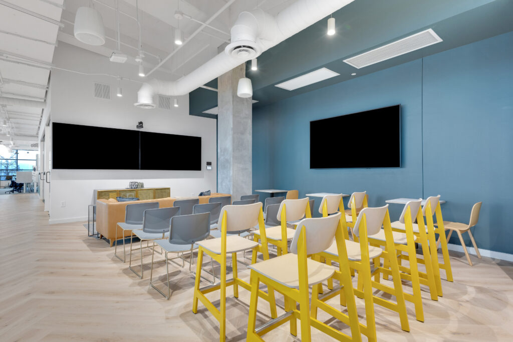 A modern office space with rows of grey and yellow chairs facing large wall-mounted monitors.
