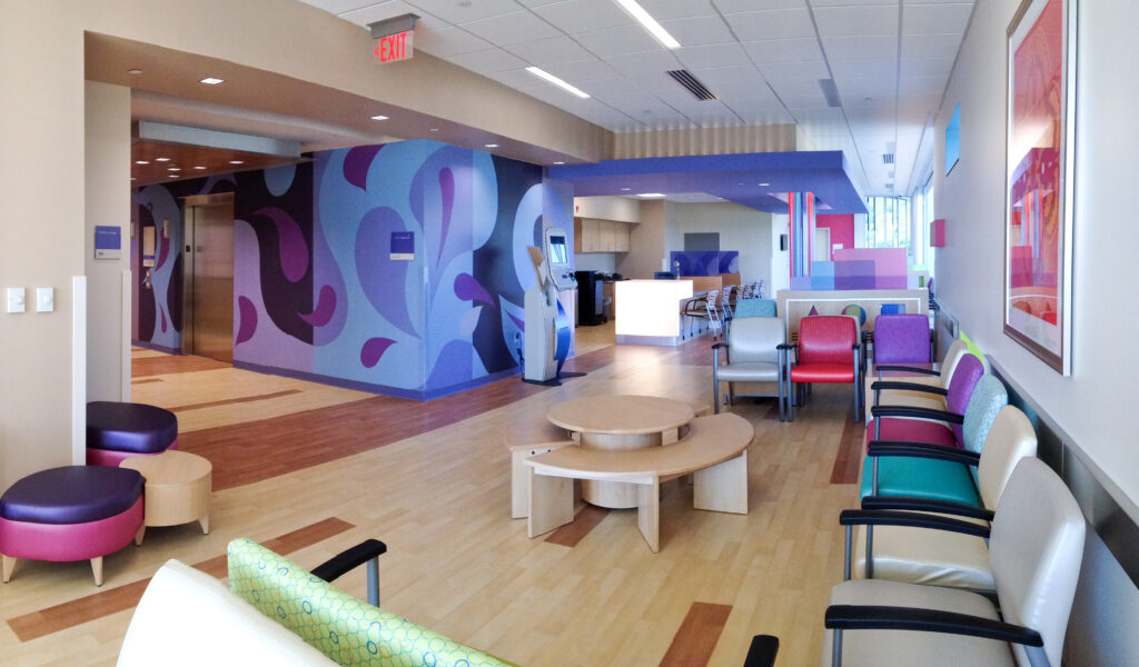Waiting area of a medical facility with a purple patterned wall, a self-service check-in kiosk, circular tables, and rows of colorful chairs.