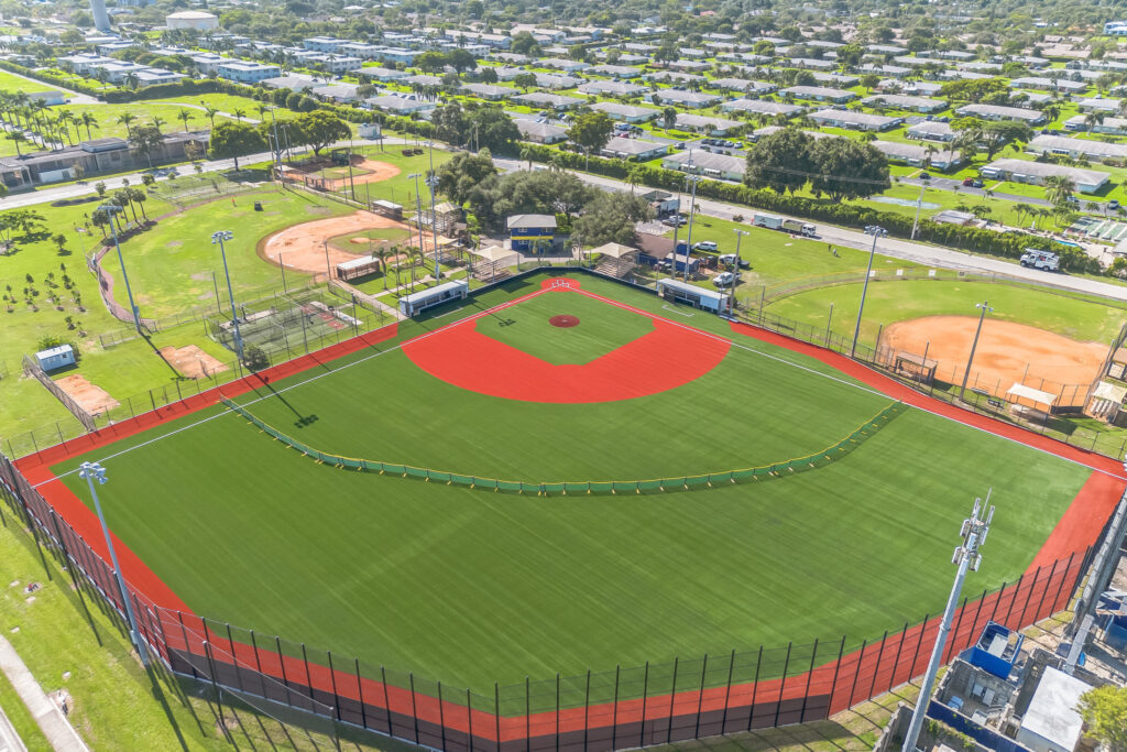 Aerial view of a baseball complex featuring a large field with green artificial turf and a red dirt infield, surrounded by a residential neighborhood.