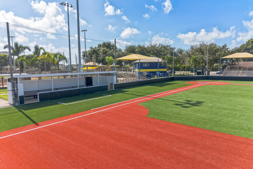 An outdoor baseball field with an artificial turf infield, dugout, and bleachers under a clear blue sky.