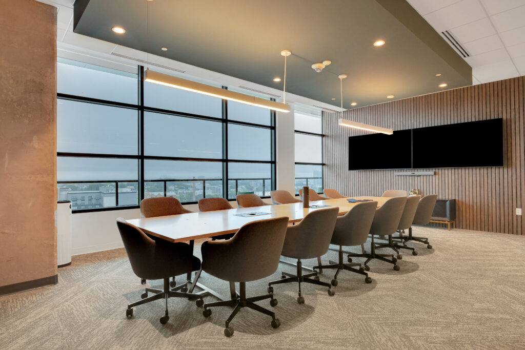 A long conference table with grey office chairs in a room with floor-to-ceiling windows, two pendant lights, and a large wall-mounted monitor.