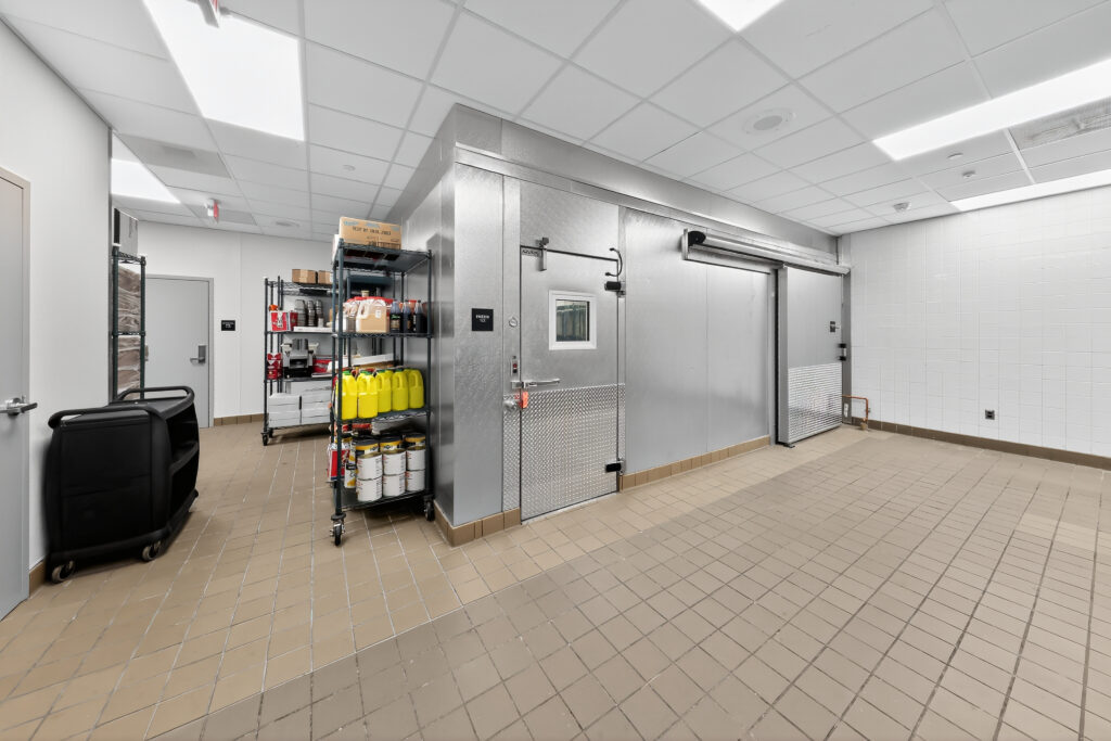 A metal walk-in cooler with sliding and swinging doors, next to a metal shelving unit stocked with cleaning supplies and containers in a tiled commercial kitchen.