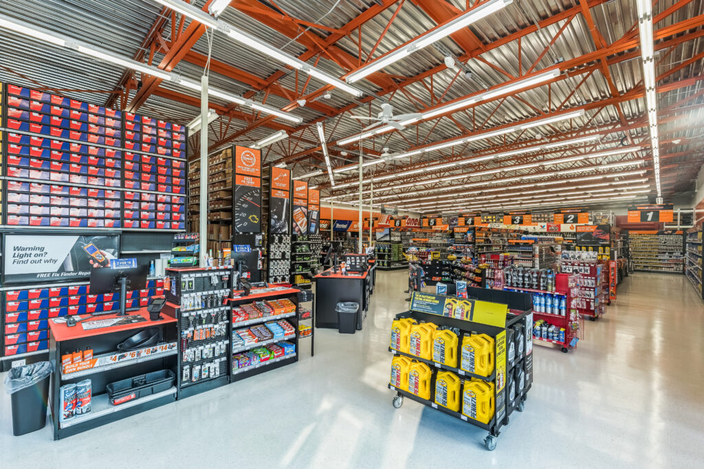 Interior of an auto parts store with aisles of shelves, wall-mounted product bins, and a display of yellow motor oil jugs in the foreground.