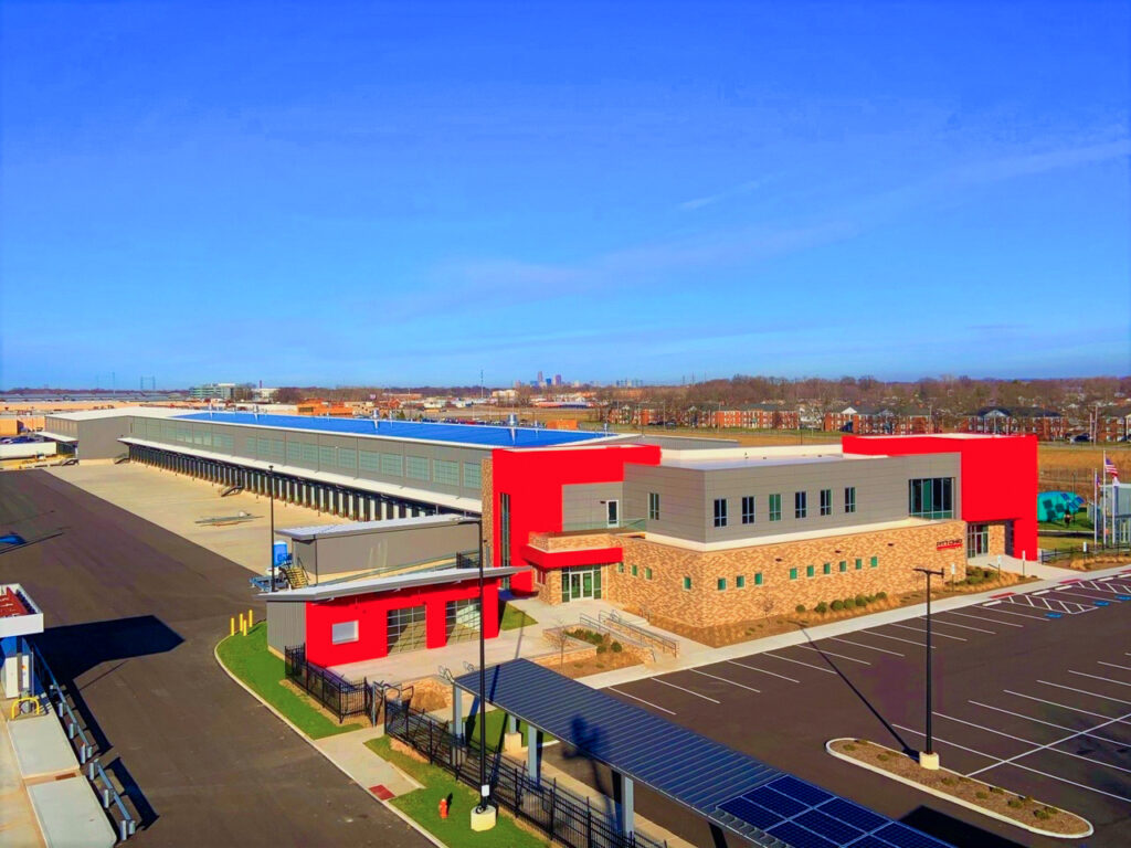 Aerial view of a large warehouse building with a red and gray facade, an expansive loading dock, and a parking lot.