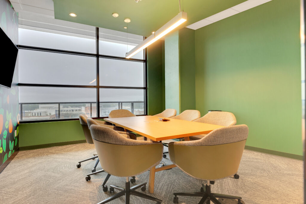 A conference room with a wooden table surrounded by six upholstered swivel chairs, situated against a green wall and a large window overlooking a city.