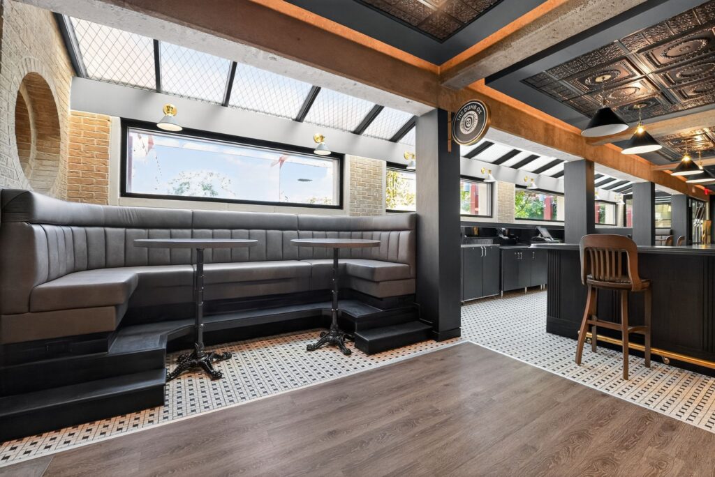 Interior of a restaurant with a grey wrap-around booth, small round tables, and a bar area with wooden stools.