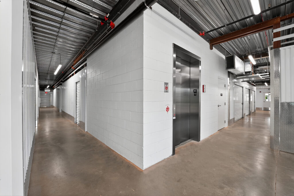 A hallway in a storage facility with a stainless steel elevator door at the corner of a white brick wall.