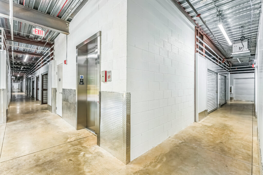 An elevator and a corridor of storage units in a building with exposed metal ceilings and concrete floors.