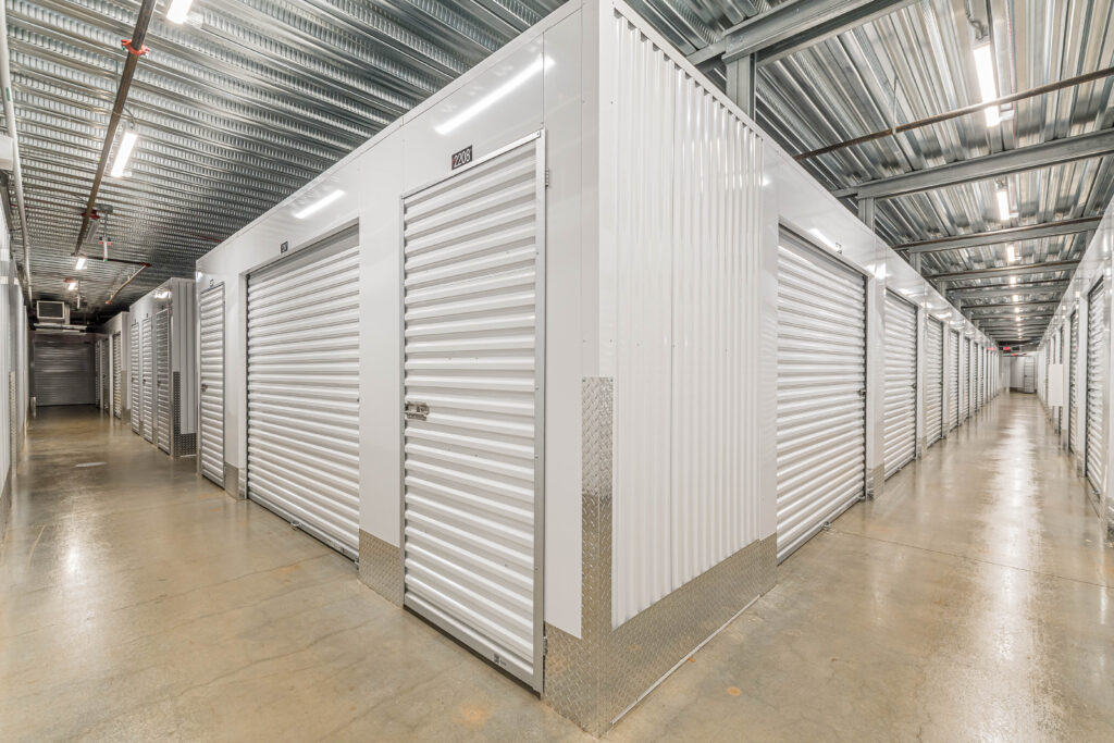 Indoor self-storage facility with a long row of white metal storage units under a corrugated steel ceiling.