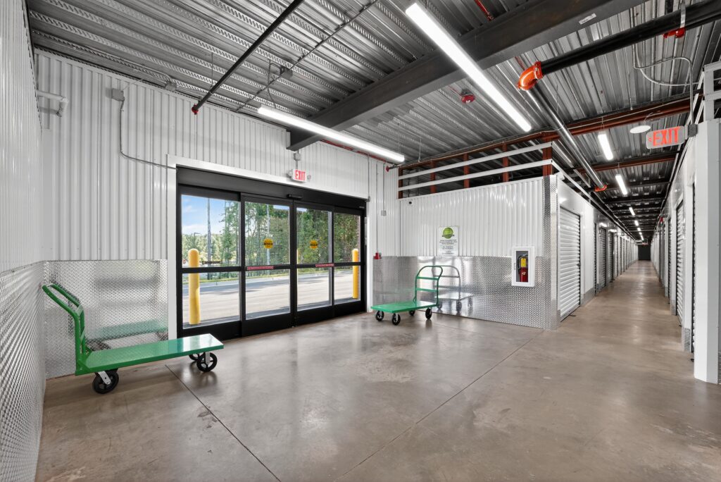 An interior view of a self-storage facility hallway featuring two flatbed carts and a glass sliding door exit.