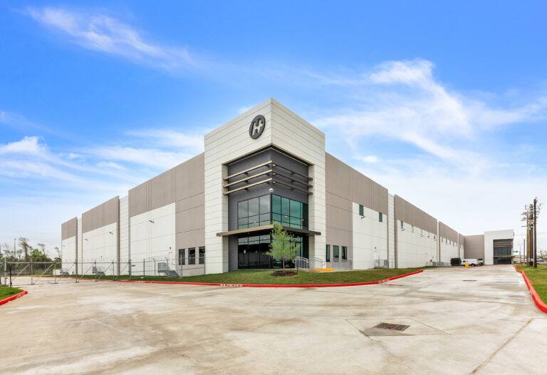 A large, modern warehouse building with a prominent “H” logo on the corner facade and a concrete parking lot in the foreground.