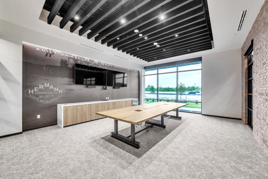 Modern office conference room with a long wood-top table, wall-mounted television, and a branded wall mural featuring the Hermann Forwarding Co. logo.