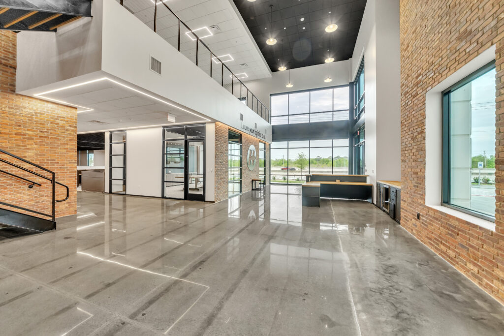 Modern office lobby with polished concrete floors, exposed brick walls, and a multi-level reception area featuring large windows.