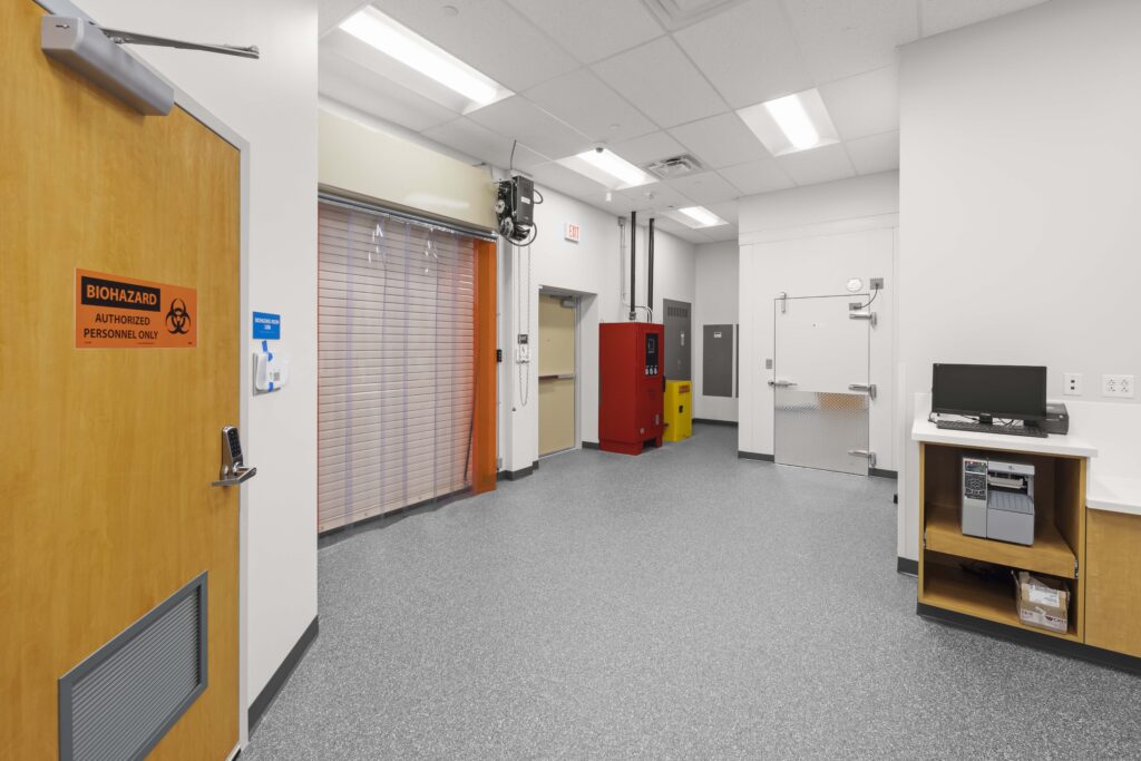 Interior of a laboratory with a biohazard door, an industrial roll-up door, a red safety cabinet, and a workstation with a printer.