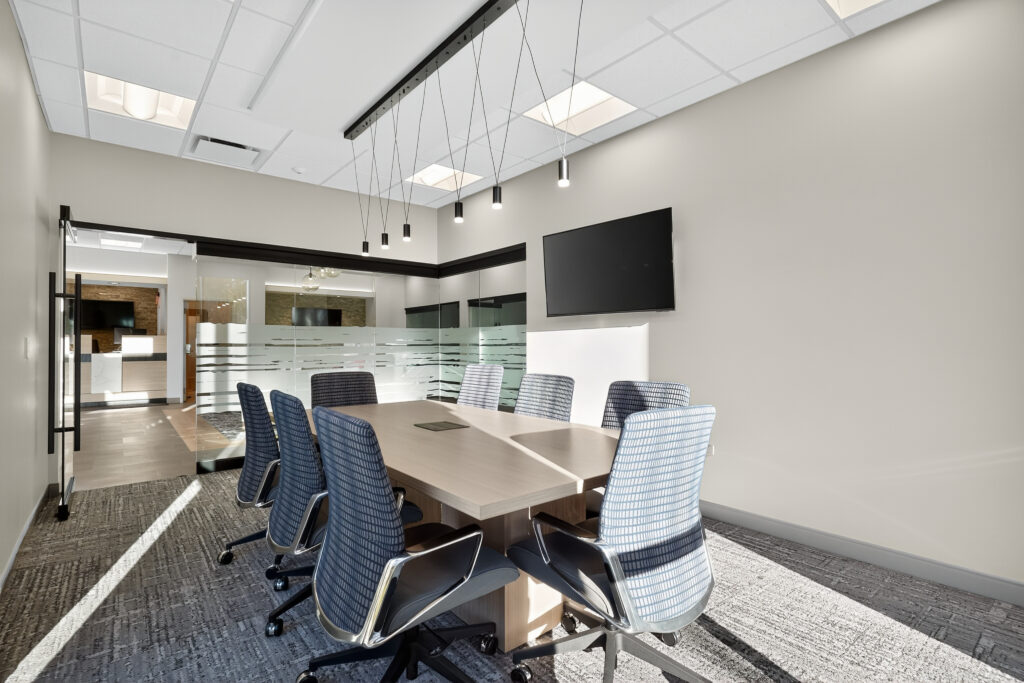 A modern conference room with a wooden table, six blue rolling chairs, and a wall-mounted flat-screen television.
