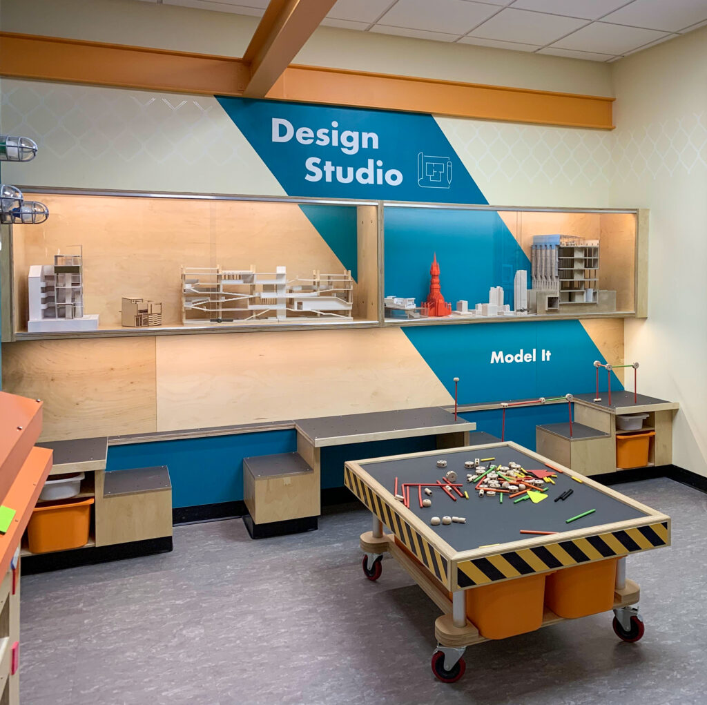 A children’s “Design Studio” area with architectural models on wall shelves and a rolling activity table with building parts.