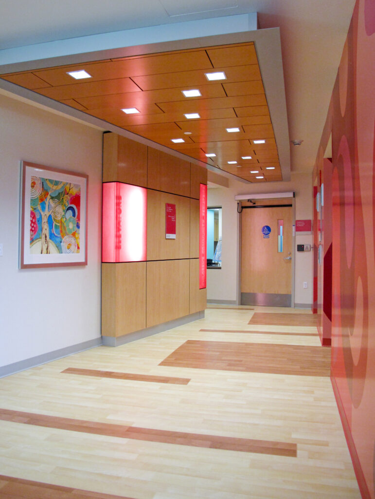 A hallway leading to an elevator bank, featuring wooden panels with red backlit signage and an abstract painting on the wall.