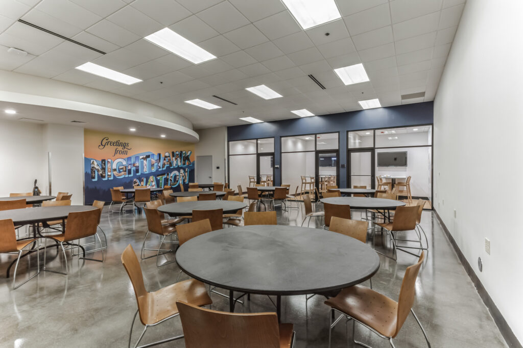 A cafeteria with several round tables, wooden chairs, and a wall mural that reads “Greetings from NIGHTHAWK NATION.”
