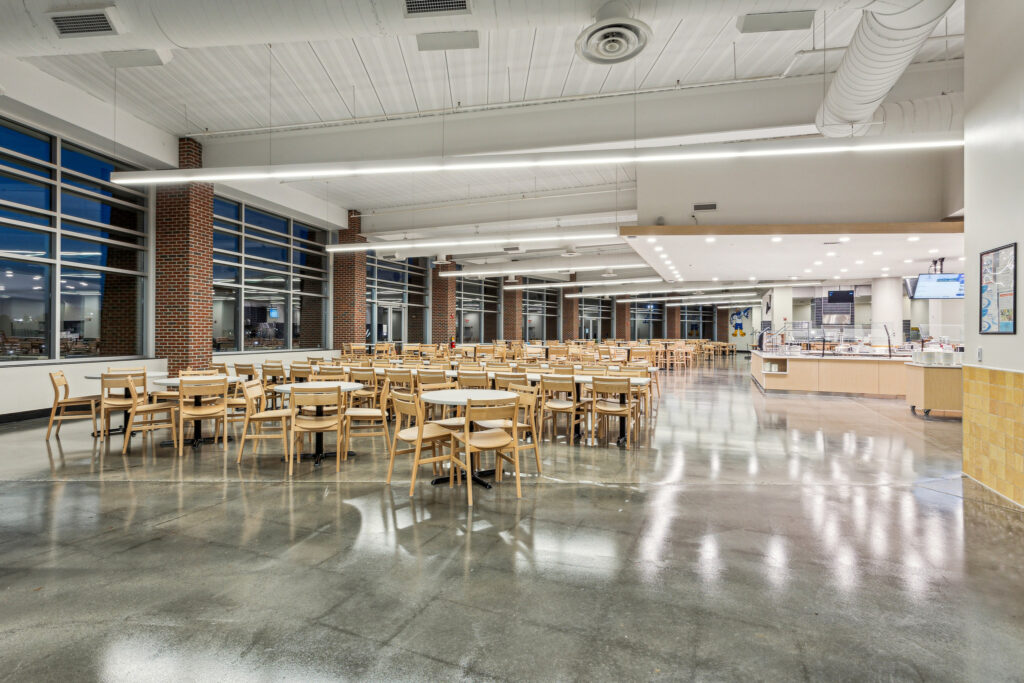 A spacious dining hall with rows of wooden chairs and circular tables, featuring floor-to-ceiling windows, brick pillars, and a service counter in the background.