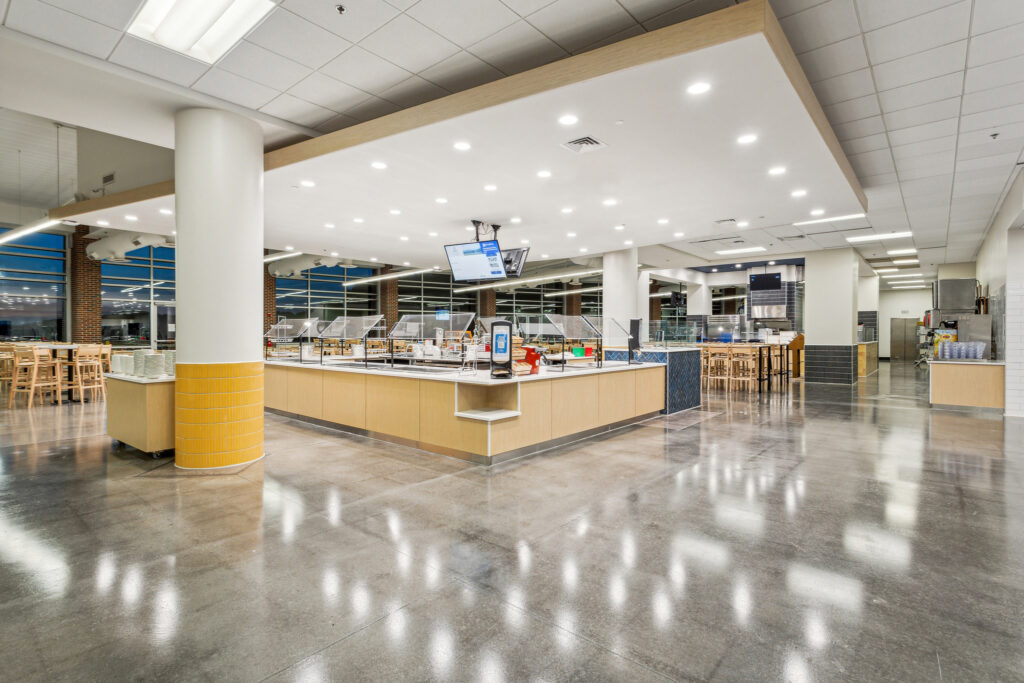 A modern, empty cafeteria featuring a central serving counter with food shields and a polished concrete floor.
