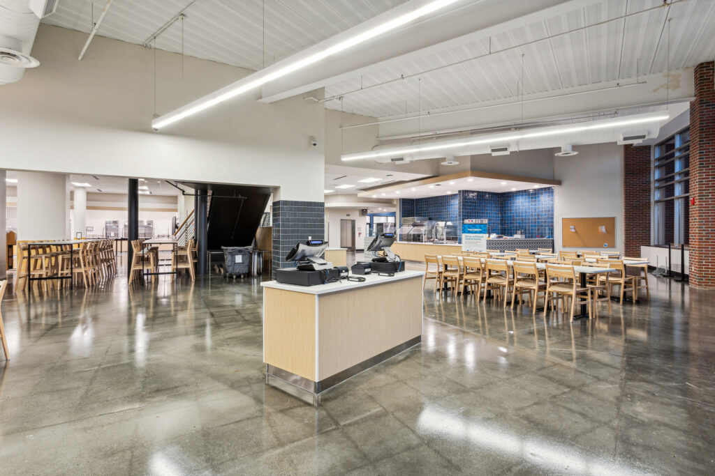 A dining hall with a central service counter, rows of tables and chairs, and a cafeteria-style food serving area in the back.