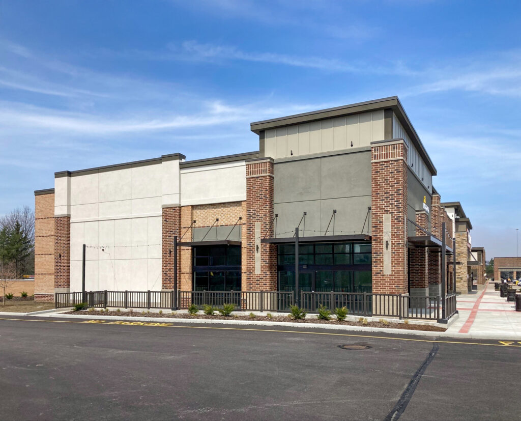 A modern commercial building with brick and stucco exterior, large glass storefront windows, and a metal railing along the front patio.