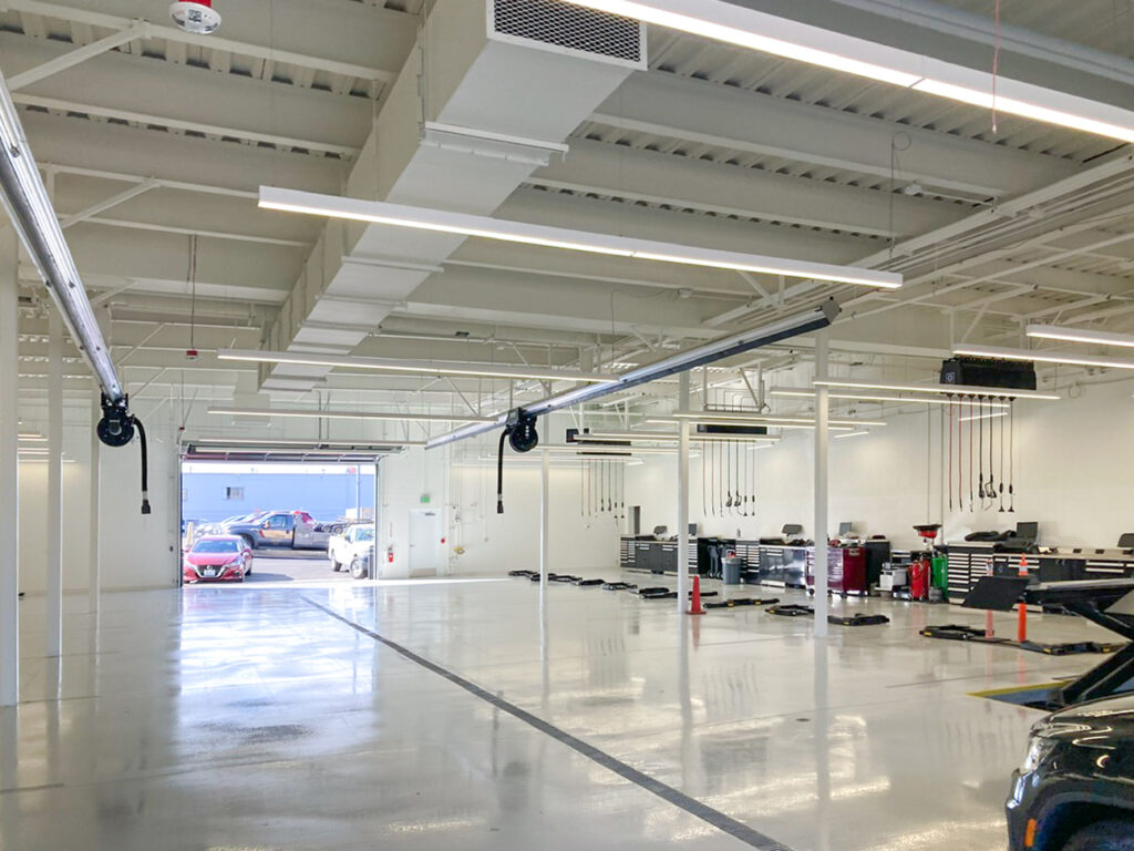 An empty, modern vehicle service bay with high ceilings, overhead exhaust hoses, and a car parked outside the open bay door.