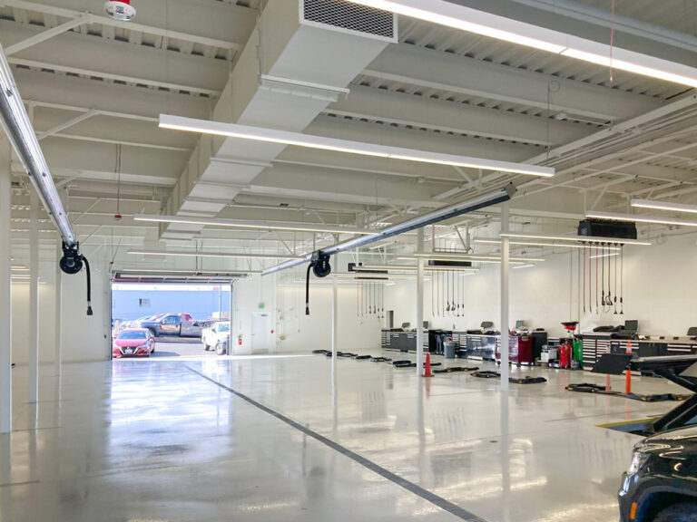 An empty, modern vehicle service bay with high ceilings, overhead exhaust hoses, and a car parked outside the open bay door.