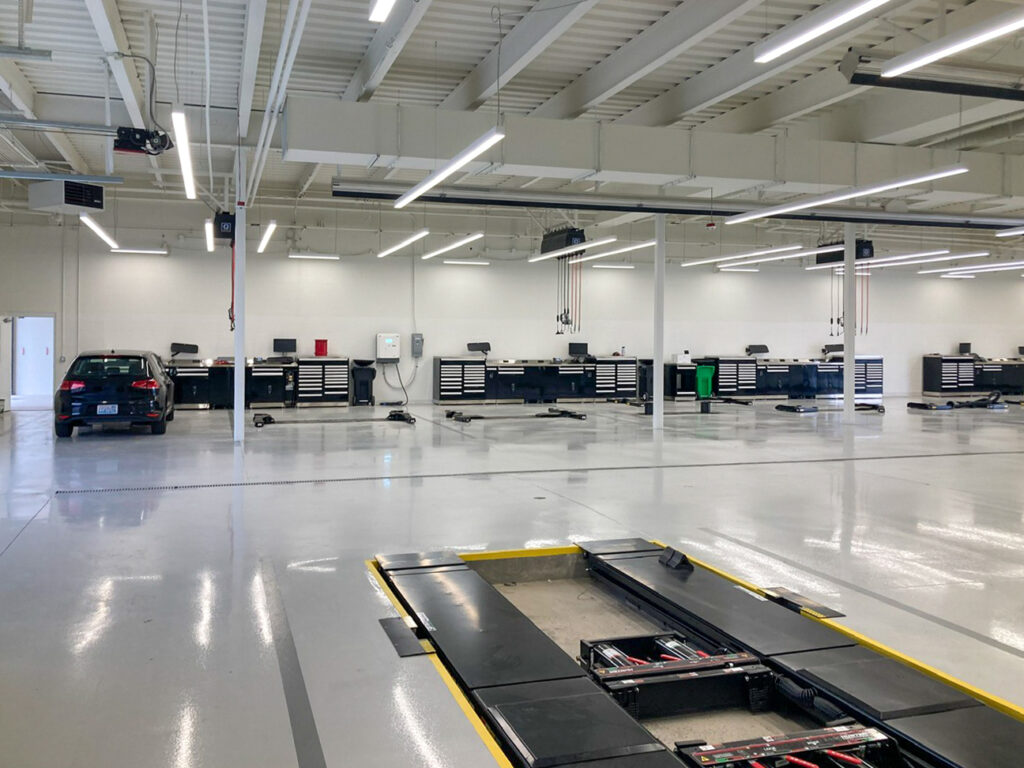 An empty vehicle service center with a car parked on the left, rows of workbenches along the back wall, and an in-ground vehicle lift in the foreground.