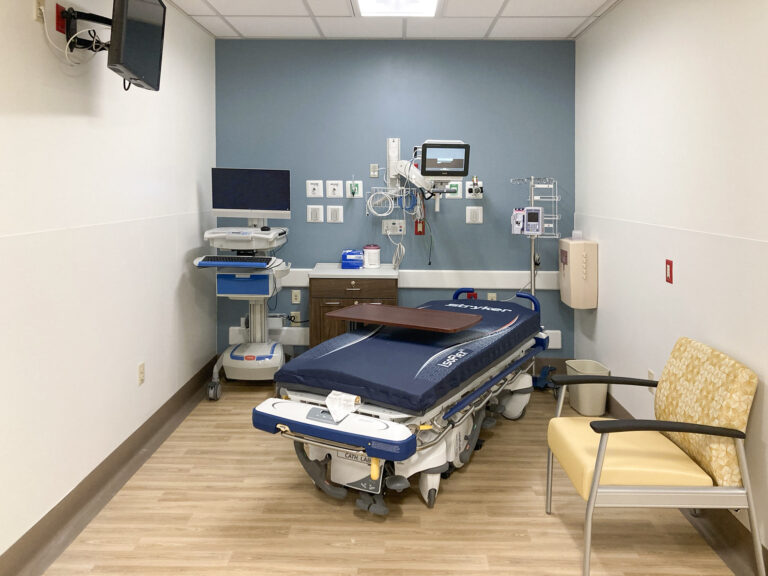 A hospital room featuring a Stryker procedure table in the center, a computer workstation on the left, and a single armchair on the right.