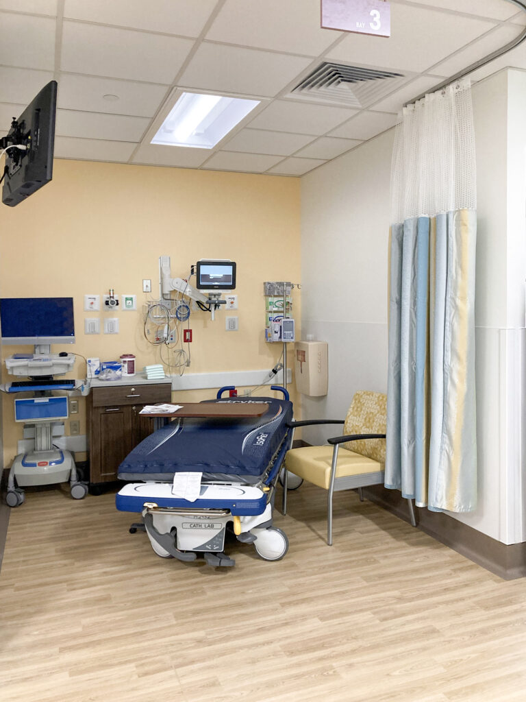 A hospital patient bay with a medical bed, monitors, medical equipment, and a chair.