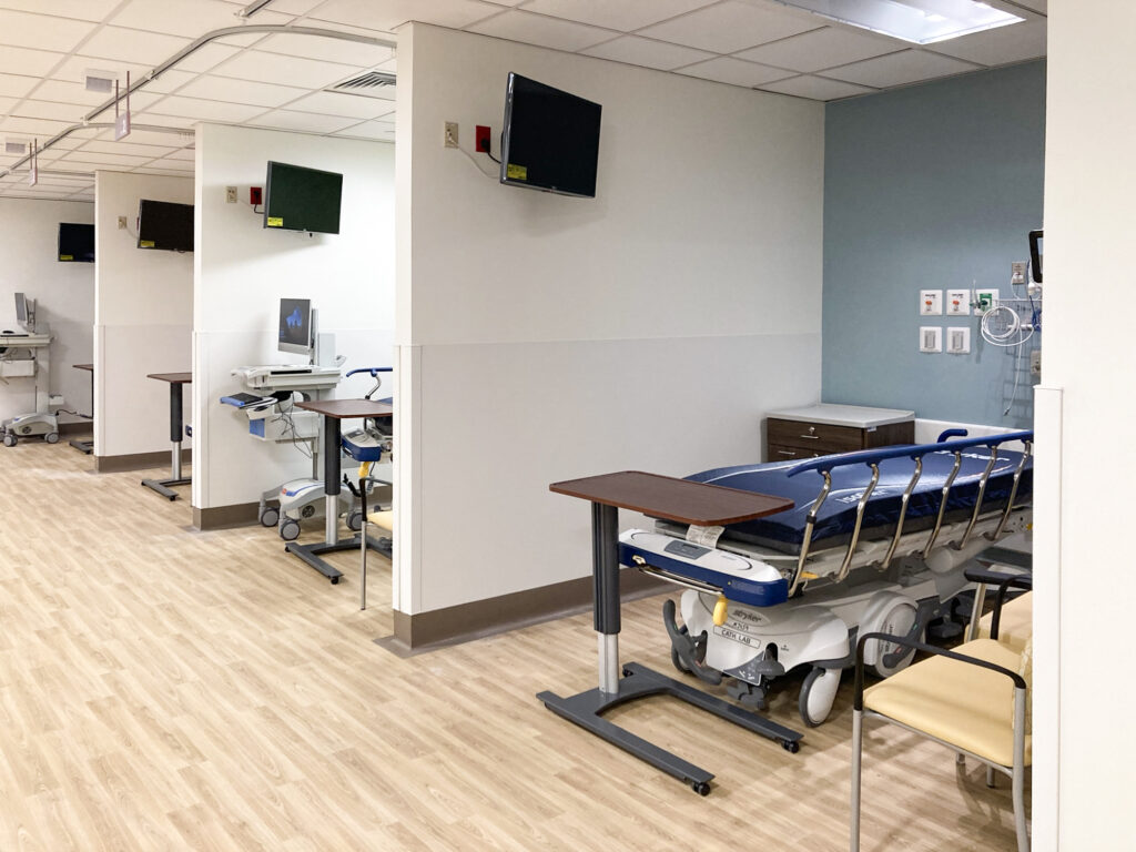 A row of hospital bays, each containing a medical gurney, an adjustable bedside table, and a wall-mounted monitor.